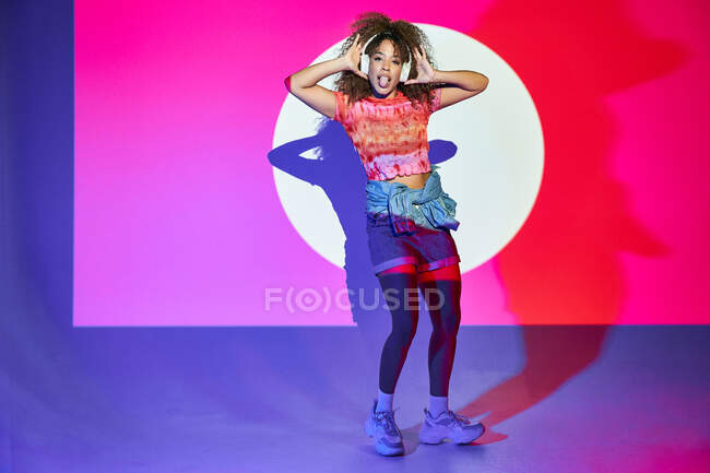 Full body African American female in shorts listening to favorite music in headphones and dancing while looking at camera making faces with tongue out in neon lights in studio — Stock Photo