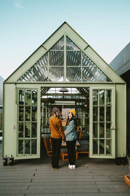 Vista trasera de una alegre pareja de hipsters de moda parados en una terraza de madera mientras se toman de la mano y miran por encima del hombro - foto de stock