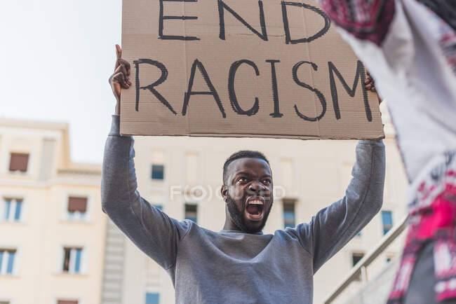 Low angle of African American male activist with End Racism poster screaming on city street during Black Lives Matter protest — Stock Photo