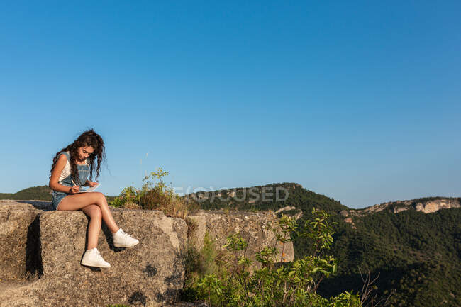 Viajante do sexo feminino sentado na rocha em terras altas e escrevendo em caderno durante trekking no verão — Fotografia de Stock