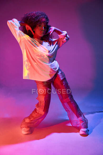 Joven bailarina afroamericana en traje informal suelto tocando el pelo rizado y mirando a la cámara mientras baila en un estudio oscuro con luces de neón - foto de stock