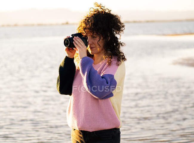 Молодая кудрявая этническая женщина-путешественница фотографирует на фотокамеру, стоя у моря в солнечный вечер — стоковое фото