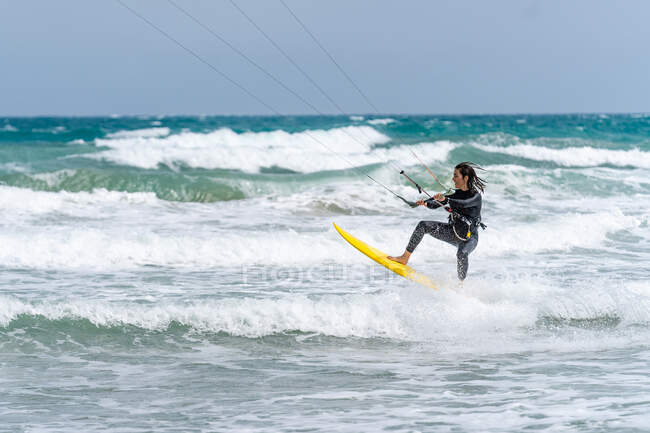 Atleta feminina ativa no kiteboard segurando barra de controle enquanto pratica kitesurf e olhando para o oceano espumoso — Fotografia de Stock