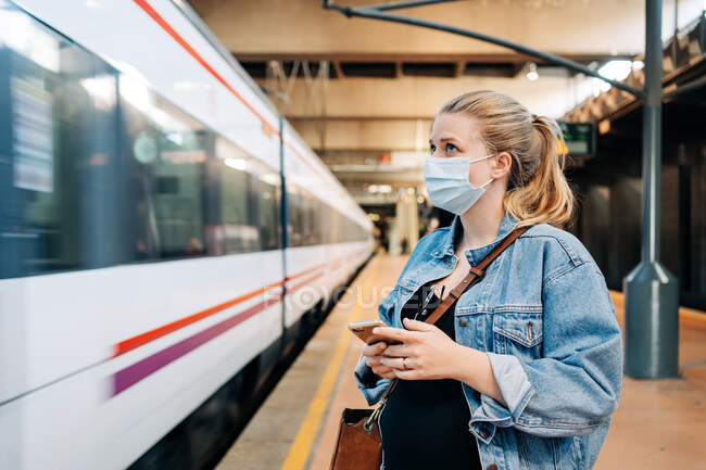 Giovane donna in abiti casual e maschera protettiva in piedi sulla stazione ferroviaria e guardando lontano in attesa di treno — Foto stock