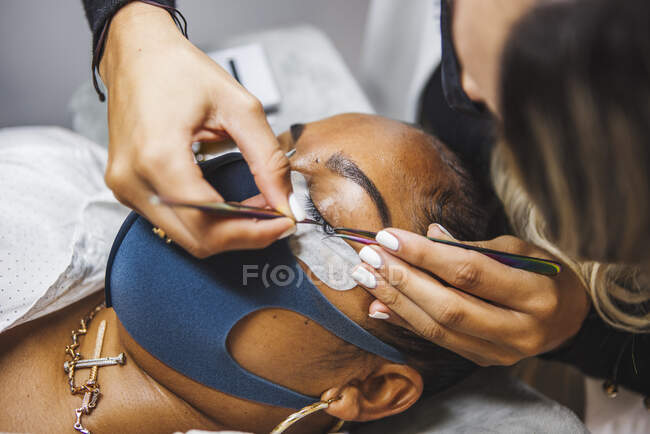 High angle of crop unrecognizable cosmetologist with tweezers applying fake eyelashes for extension on eye of ethnic client with face protective mask in salon during coronavirus pandemic — Fotografia de Stock