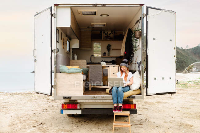 Viaggiatore femminile deliziato seduto fuori sulle scale del furgone mentre si lavora in remoto sul computer portatile — Foto stock