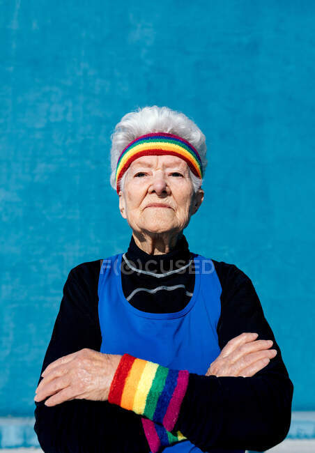 Крутая пожилая женщина в повязке и браслетах, стоящая со скрещенными руками на синем фоне — стоковое фото