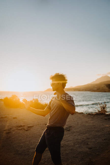 Vista laterale viaggiatore maschile in abiti casual e occhiali da sole battendo le mani sulla costa sabbiosa al pittoresco tramonto mentre trascorre le vacanze estive in Fyriplaka Grecia — Foto stock