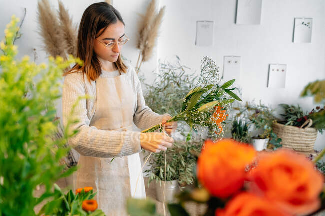 Konzentrierte junge ethnische Floristin in lässiger Kleidung und Schürze, die einen zarten Strauß orangefarbener Lilien und weißer Zigeunerblumen in einem Blumenladen bindet — Stockfoto