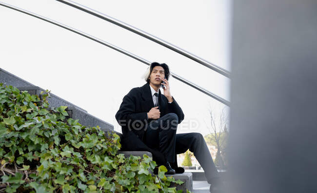 Bajo ángulo de joven empresario asiático bien vestido conversando en el teléfono celular mientras está sentado en la escalera urbana y mirando hacia otro lado - foto de stock