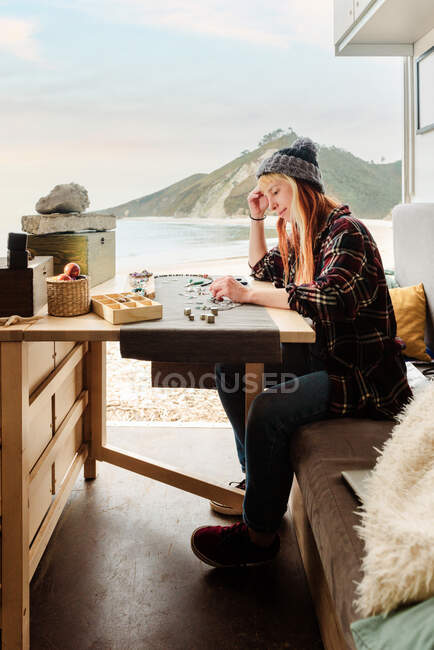 Vista lateral del viajero femenino enfocado creando accesorios hechos a mano mientras está sentado en la mesa de madera en un camión estacionado en la playa - foto de stock