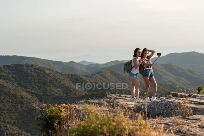 Подорожуючі друзі-жінки з рюкзаками, що стоять на пагорбі і роблять самостріл на смартфоні на тлі гірського хребта влітку — стокове фото