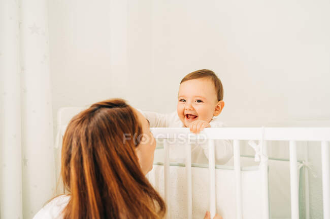 Мама сидит рядом с очаровательным радостным ребенком в белой колыбели в светлой детской комнате — стоковое фото