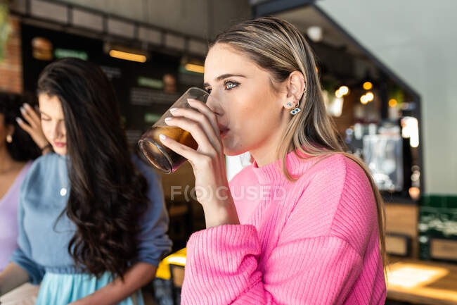 Vista lateral de la joven hembra de moda en traje de beber vaso de coca fría y mirando hacia otro lado mientras pasa tiempo con amigos en la cafetería - foto de stock