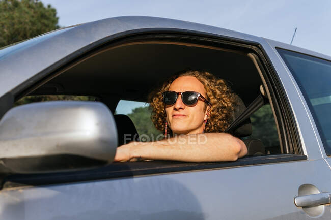 Ernster junger Mann mit stylischer Sonnenbrille blickt durch das geöffnete Autofenster in die Kamera, während er am Fahrersitz sitzt — Stockfoto