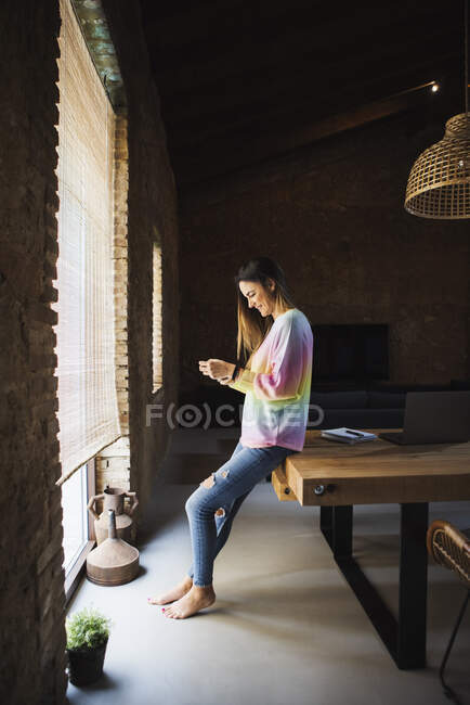 Side view of cheerful female surfing internet on cellphone while sitting on table in loft style house - foto de stock