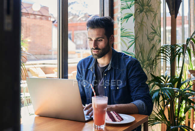 Side view of ethnic male employee working on netbook at table with tasty cake piece and drinks — Stock Photo