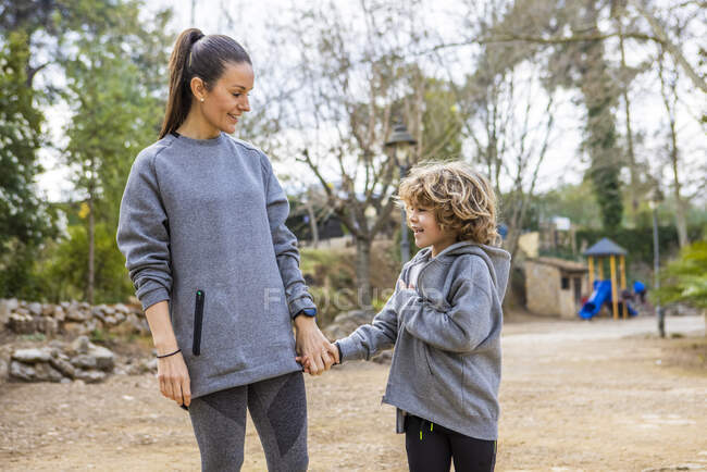 Mother in sportswear holding boy by hand while strolling on walkway and talking against trees looking at each other - foto de stock