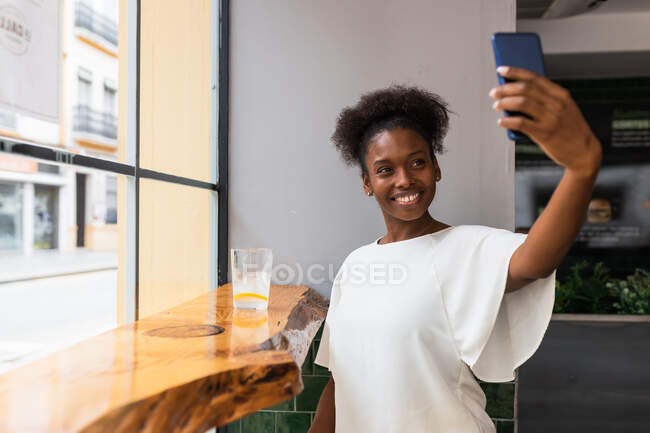 Jovem afro-americana feliz em blusa branca tirando selfie no celular enquanto se senta na mesa alta perto da parede de vidro no café moderno — Fotografia de Stock