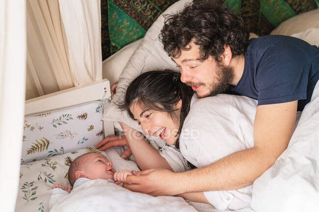 Сверху веселые родители, взаимодействующие с милым маленьким ребенком, лежа в постели дома — стоковое фото