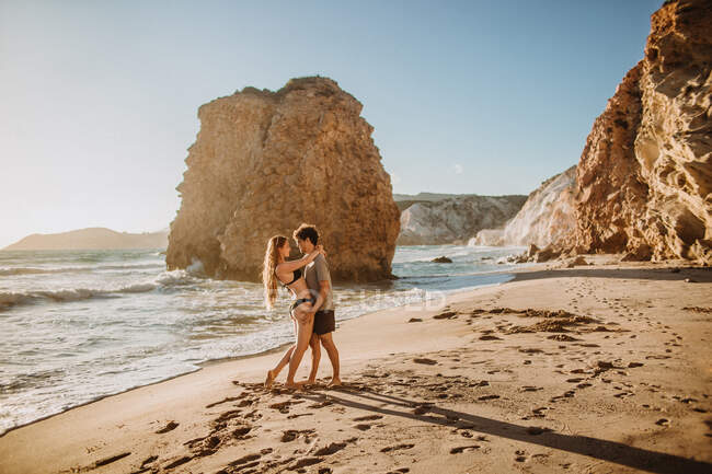 Полнотелый мужчина обнимает молодую привлекательную девушку в купальниках, расслабляясь вместе на песчаном побережье у грубых скал в солнечный день в Фириплака Милош — стоковое фото