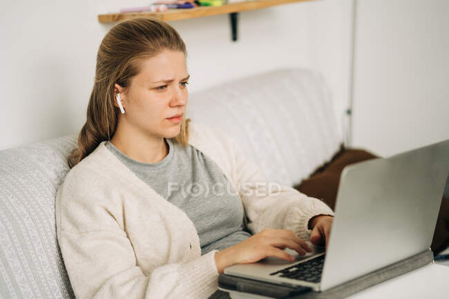 Attentive young female distance employee in earbuds typing on netbook while sitting on sofa in house room — Stock Photo