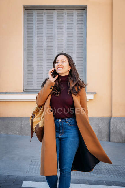 Side view of content female in coat with backpack speaking on cellphone while strolling on crosswalk and looking away — Stock Photo