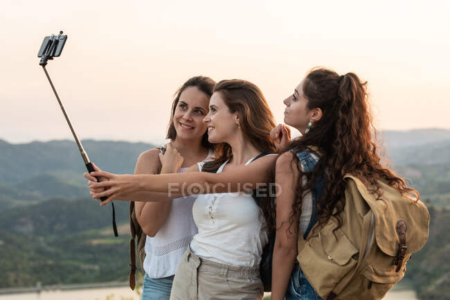 Viajando amigas con mochilas de pie en la colina y tomando fotos en el teléfono inteligente en el fondo de la cordillera en verano - foto de stock