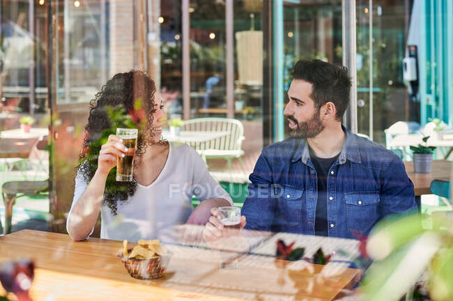 A través del vidrio alegre pareja étnica con vasos de cerveza y papas fritas hablando mientras se miran a la luz del sol - foto de stock