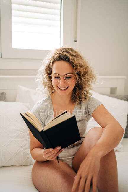 Позитивна молода жінка з кучерявим світлим волоссям в трусиках і окулярах посміхається, сидячи на затишному ліжку і читаючи цікаву книгу — стокове фото