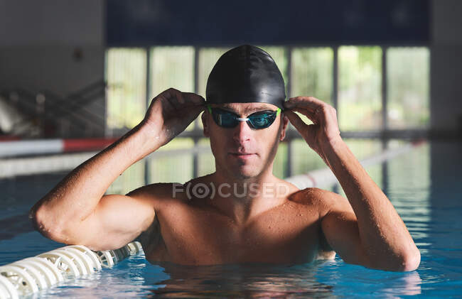 Deportista muscular adulto en gorra de natación y gafas profesionales preparándose para el entrenamiento mientras mira la cámara en la piscina con ondas - foto de stock