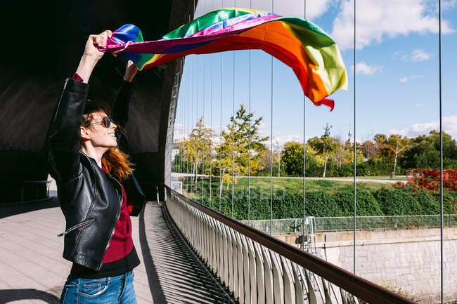 Side view of delighted lesbian female standing on bridge with LGBT rainbow flag fluttering in wind and looking away — Stock Photo