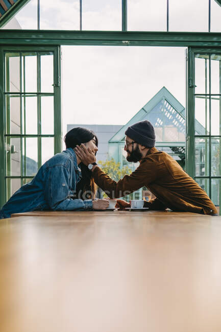 Low angle side view of tender man touching face of smiling woman while sitting at table in cafe and looking at each other with love — Stock Photo