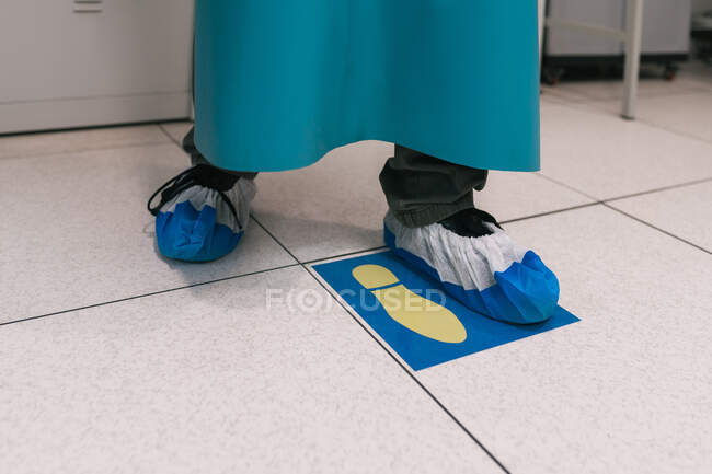 Crop anonymous chemist or doctor wearing long apron and medical overshoes standing on step floor sign in modern hospital — Stock Photo