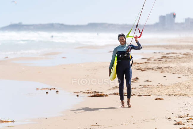 Sportlerin im Neoprenanzug mit aufblasbarem Drachen spaziert am Sandstrand und blickt in Kamera gegen stürmischen Ozean — Stockfoto
