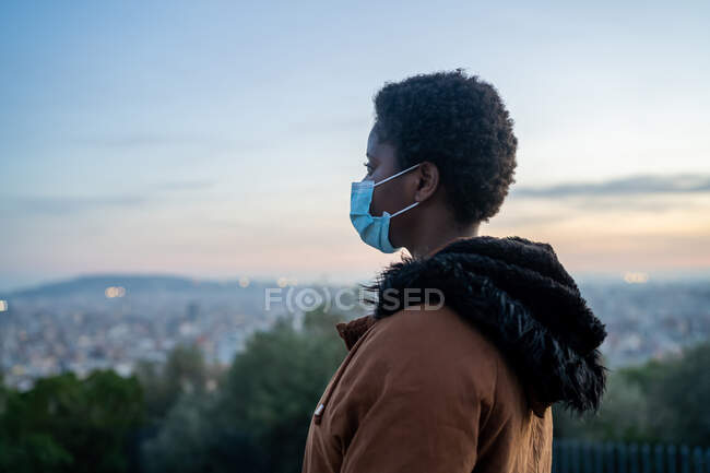 Вид сбоку на созерцательную афроамериканку в теплой куртке и маске для лица, стоящую на вершине зеленого холма и наблюдающую современный город в сумерках — стоковое фото