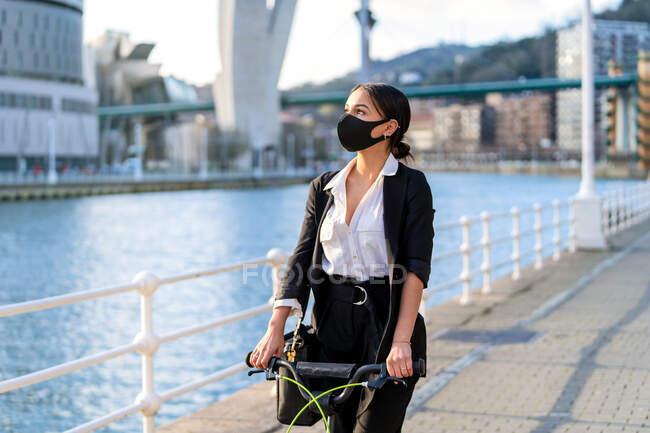 Ethnic female entrepreneur in fabric mask and formal clothes riding electric scooter on walkway in town during coronavirus pandemic looking away — Stock Photo