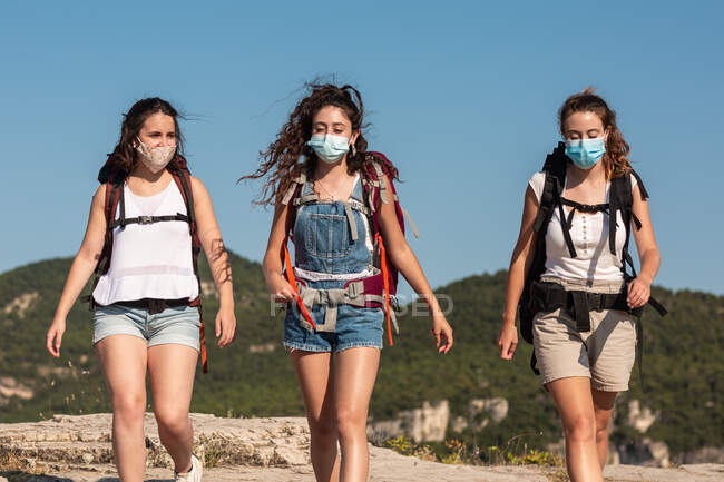 Compañía de viajeros femeninos con mochilas y en máscaras protectoras de senderismo en las tierras altas en verano - foto de stock