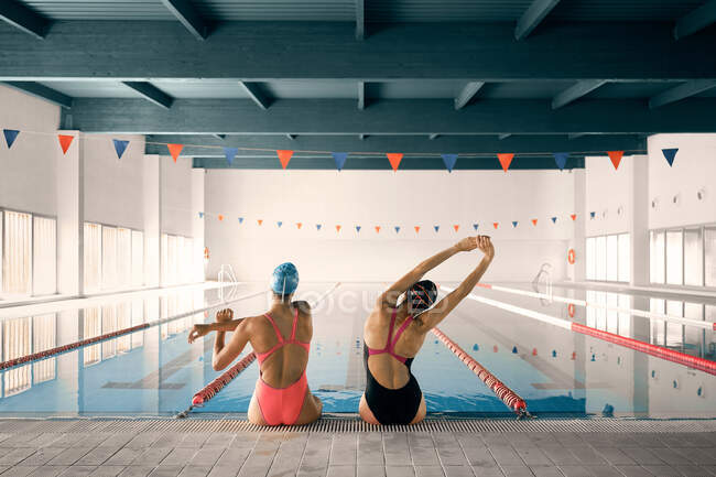 Back view of anonymous sportswomen in swimsuits stretching arms while sitting on poolside during training — Stock Photo