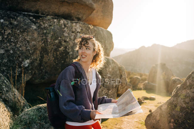 Vista lateral da jovem exploradora feliz com cabelo escuro em roupas casuais sentadas em meio a rochas rochosas e ler mapa contra o céu azul sem nuvens — Fotografia de Stock