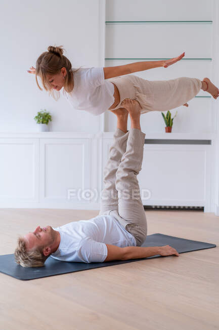 Side view of boyfriend lifting girlfriend while doing acro yoga together at home and holding hands — Fotografia de Stock