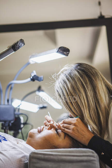 Side view of unrecognizable cosmetologist with tweezers applying fake eyelashes for extension on eye of ethnic client in salon — Fotografia de Stock