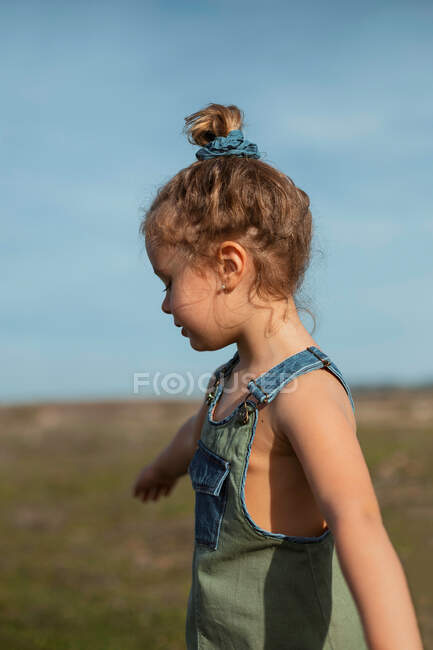 Очаровательная маленькая девочка в комбинезоне, стоящая с протянутыми руками на лугу и смотрящая вниз — стоковое фото