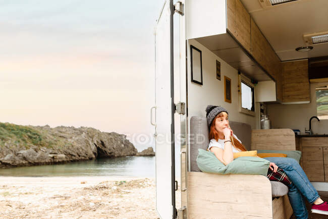 Vista laterale di riflessivo viaggiatore femminile hipster seduto sul divano in camion e godendo di vacanza vicino al lago — Foto stock