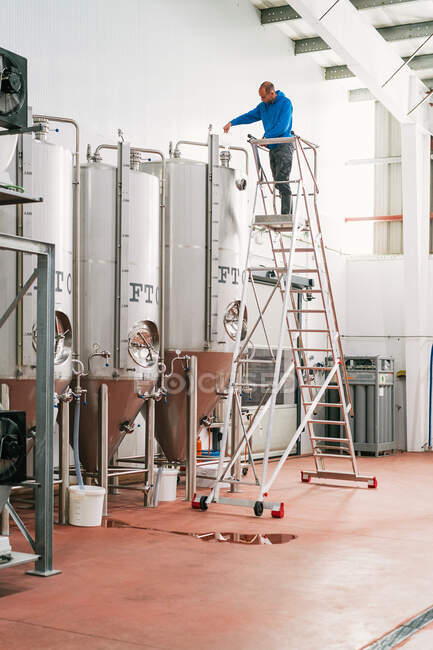 Ingegnere maschio sulla scala che versa il liquido nel serbatoio in acciaio inossidabile mentre lavora in birreria — Foto stock