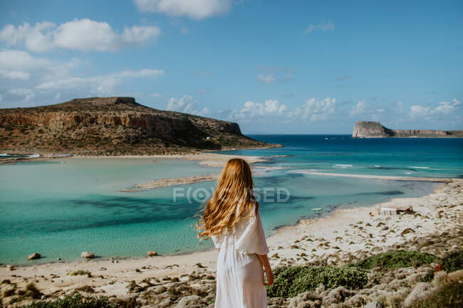 Seitenansicht anonyme Frau in weißer Maxi-Bademode, die auf einem steinigen Hügel steht und den malerischen Blick auf den Strand von Balos bewundert und an sonnigen heißen Tagen lange Haare berührt — Stockfoto
