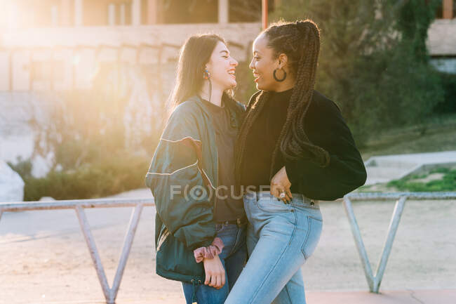 Улыбающиеся разнообразные лесбиянки в модной одежде обнимаются и разговаривают, глядя друг на друга на дорожке — стоковое фото