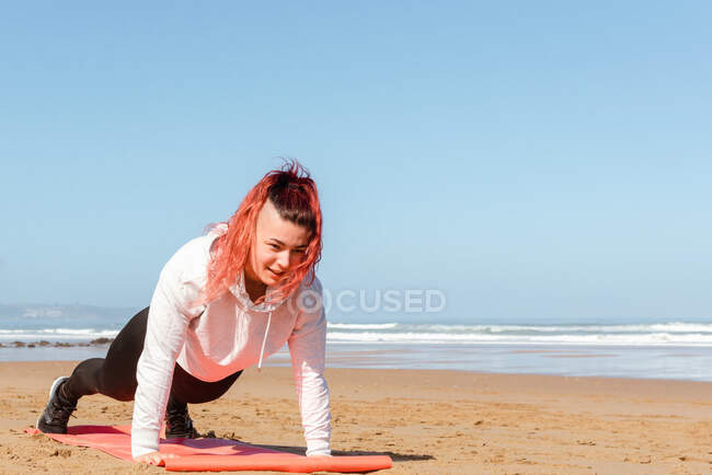Atleta sonriente con ropa deportiva mirando hacia abajo mientras hace ejercicio en la estera en la playa de arena contra el océano espumoso bajo el cielo azul - foto de stock