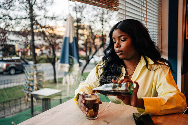 Afro-americana fêmea sentada à mesa no café e derramando refrigerante frio bebida em vidro — Fotografia de Stock