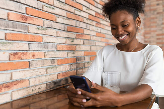 Cheerful young African American female in white shirt browsing mobile phone while sitting at table with cold drink in restaurant — Stock Photo